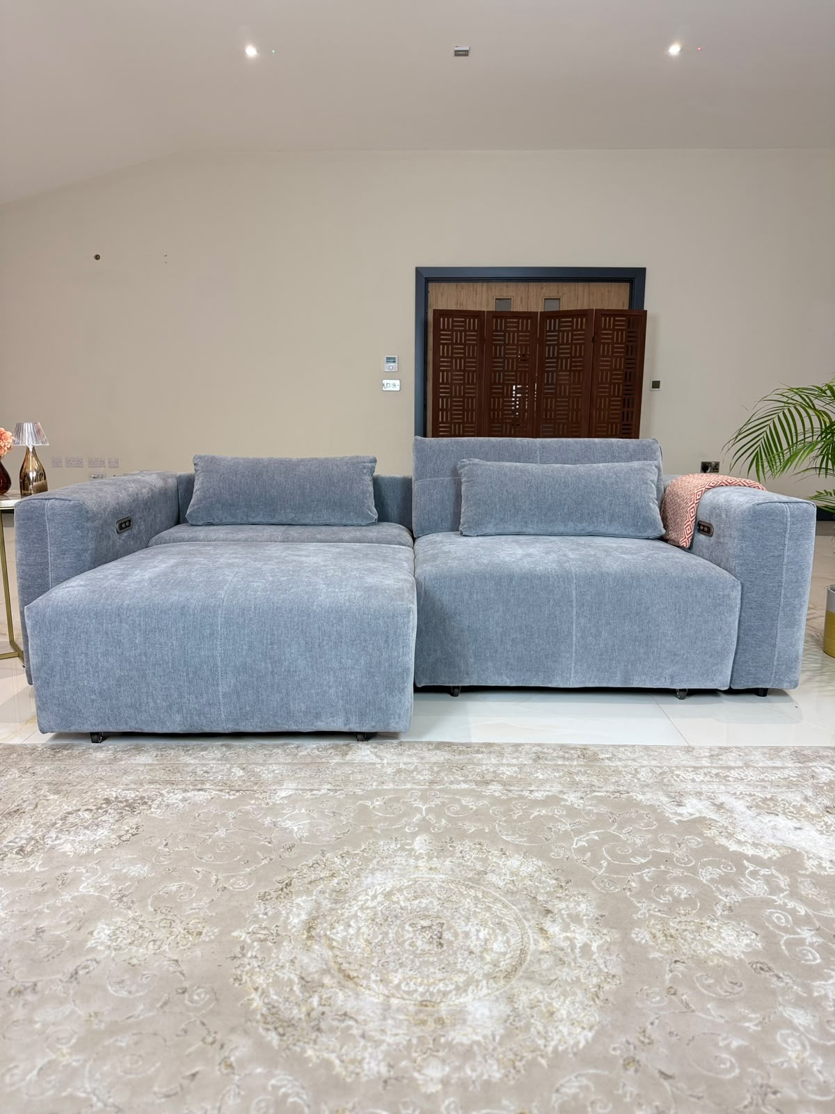 A light grey modular sectional sofa with a chaise lounge on the left, styled in a modern living room setting with a beige patterned rug and a pink accent pillow.