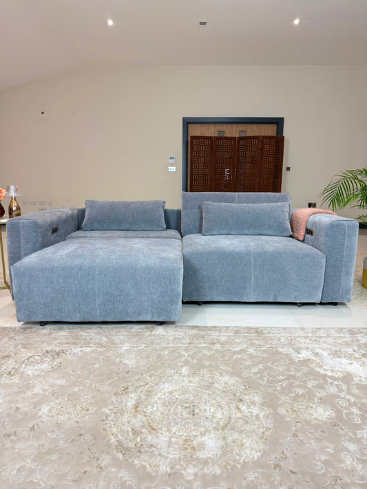 A light grey modular sectional sofa with a chaise lounge on the left, styled in a modern living room setting with a beige patterned rug and a pink accent pillow.