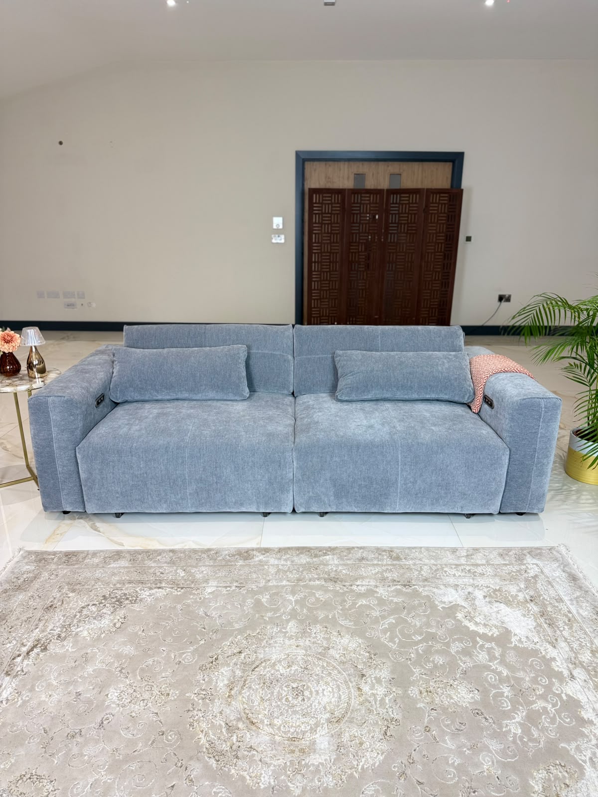 A modern light grey fabric sofa with two matching throw pillows, set in a spacious room with a beige patterned rug, a gold side table, and a potted plant.