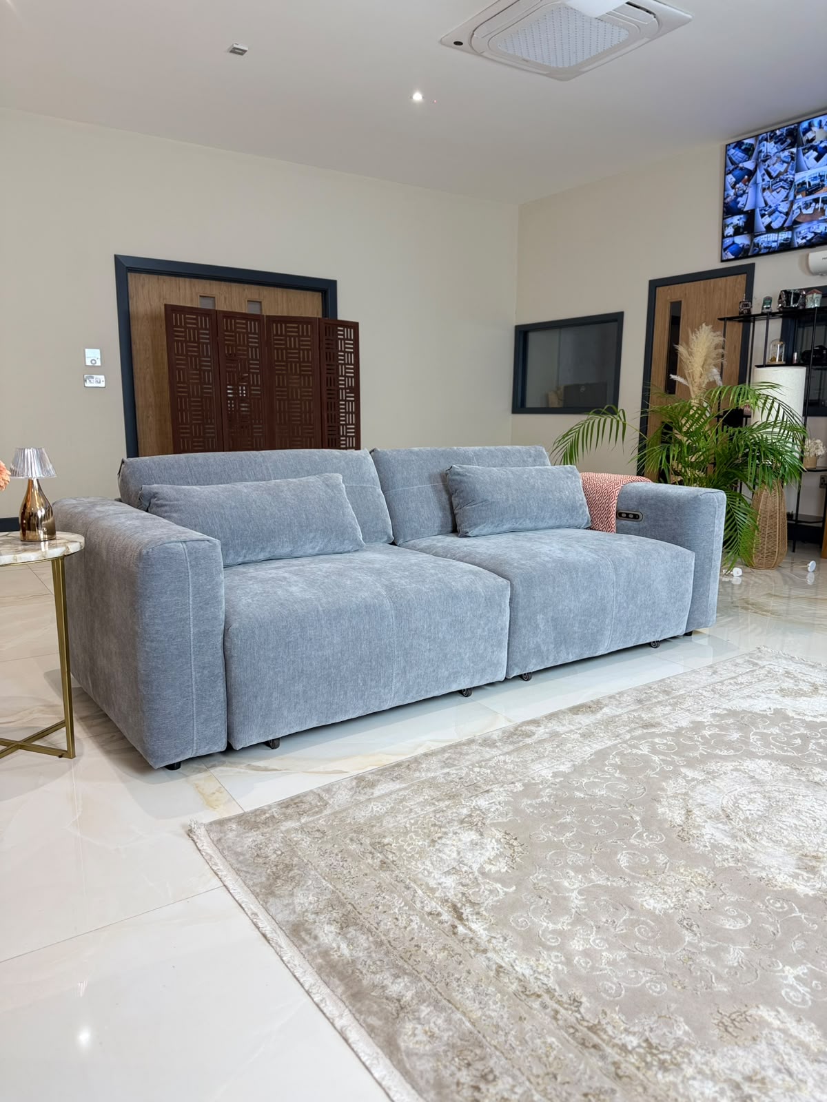 A modern light grey fabric sofa with matching cushions, styled in a contemporary living room with a beige patterned rug, a small gold side table, and indoor plants.