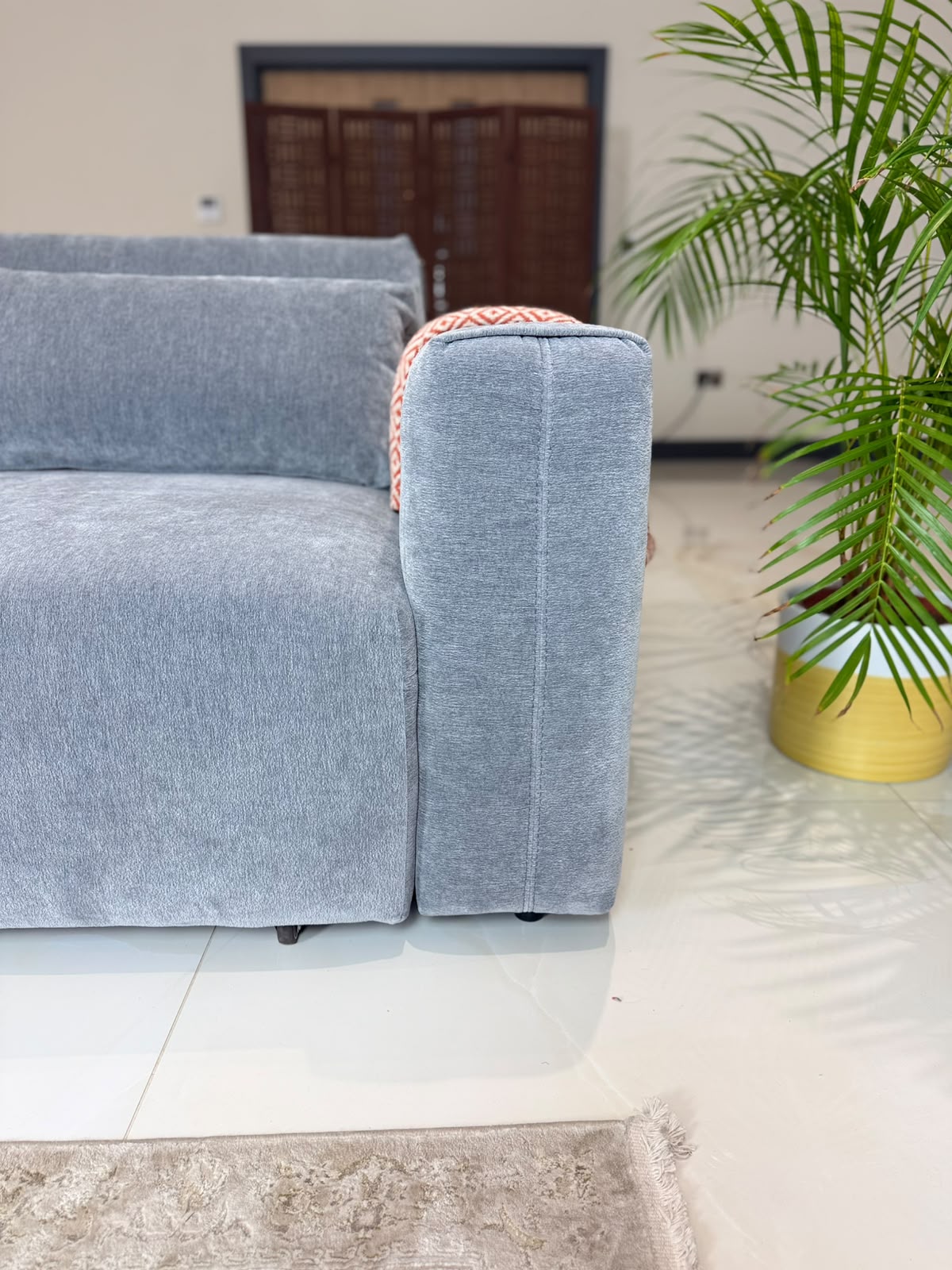 A light grey textured fabric sofa shown in a modern living room setting, with a potted green palm plant in a yellow pot on a white tiled floor.