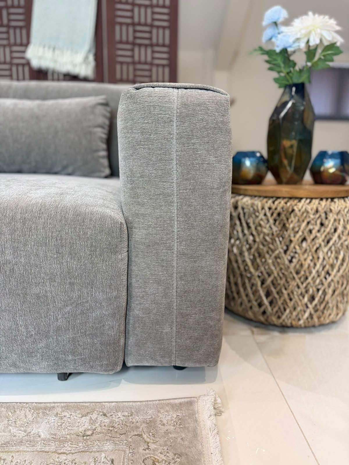 A close-up of a modern gray fabric sofa corner with a matching cushion, set in a styled living room next to a woven side table holding a vase of flowers.