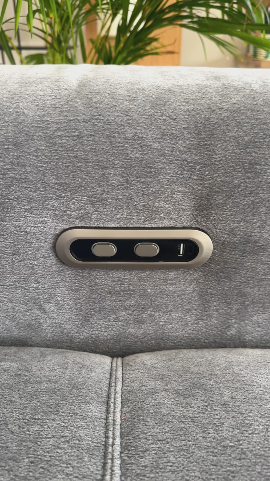 Close-up of a gray upholstered chair or sofa back with a recessed control panel featuring three buttons and a small USB-C port, with blurred green plants in the background.