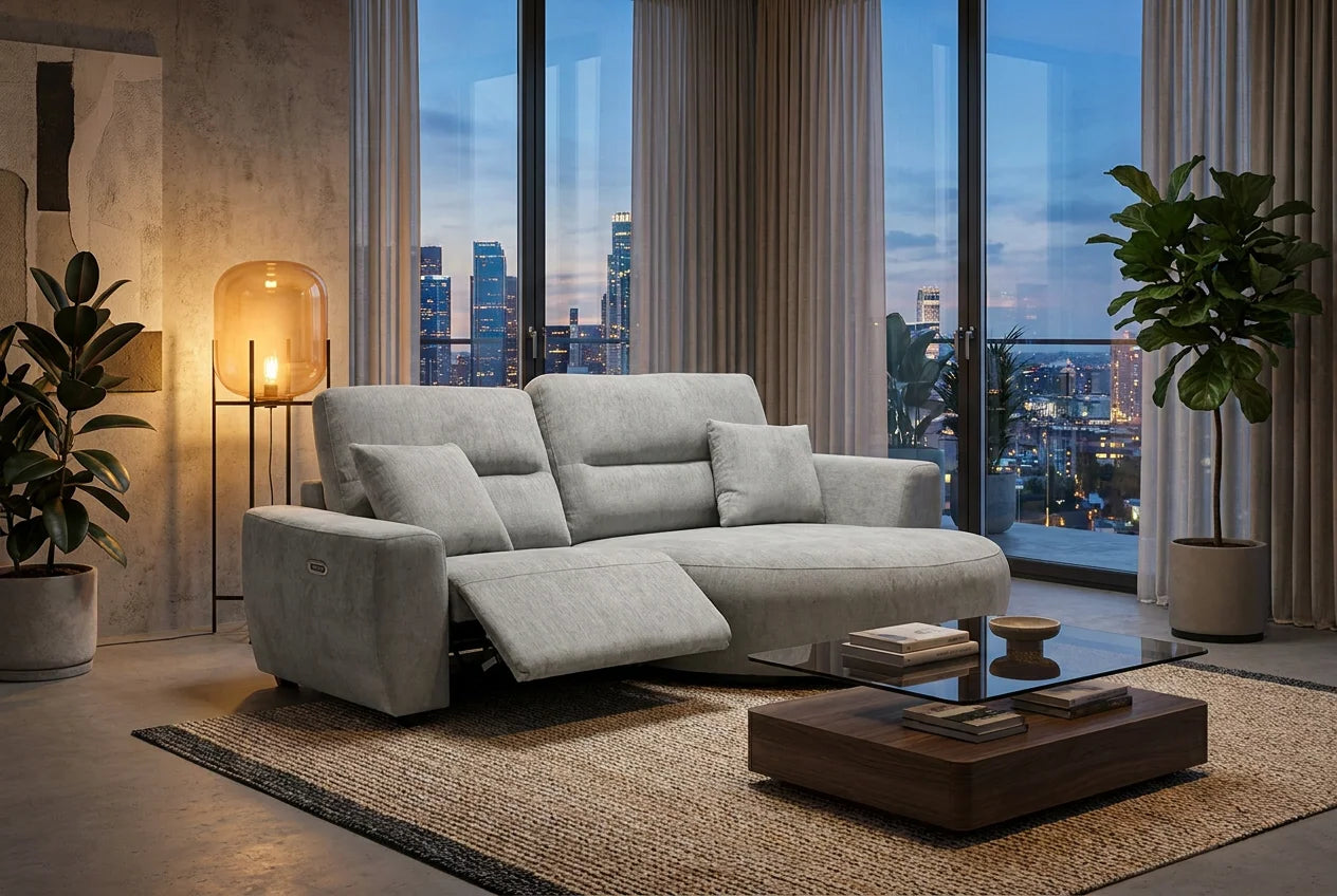 A light gray upholstered reclining sofa in a modern living room with large floor-to-ceiling windows overlooking a city skyline at dusk. The sofa is shown with the chaise extended, placed on a woven rug beside a dark wood coffee table, with plants and a floor lamp nearby.