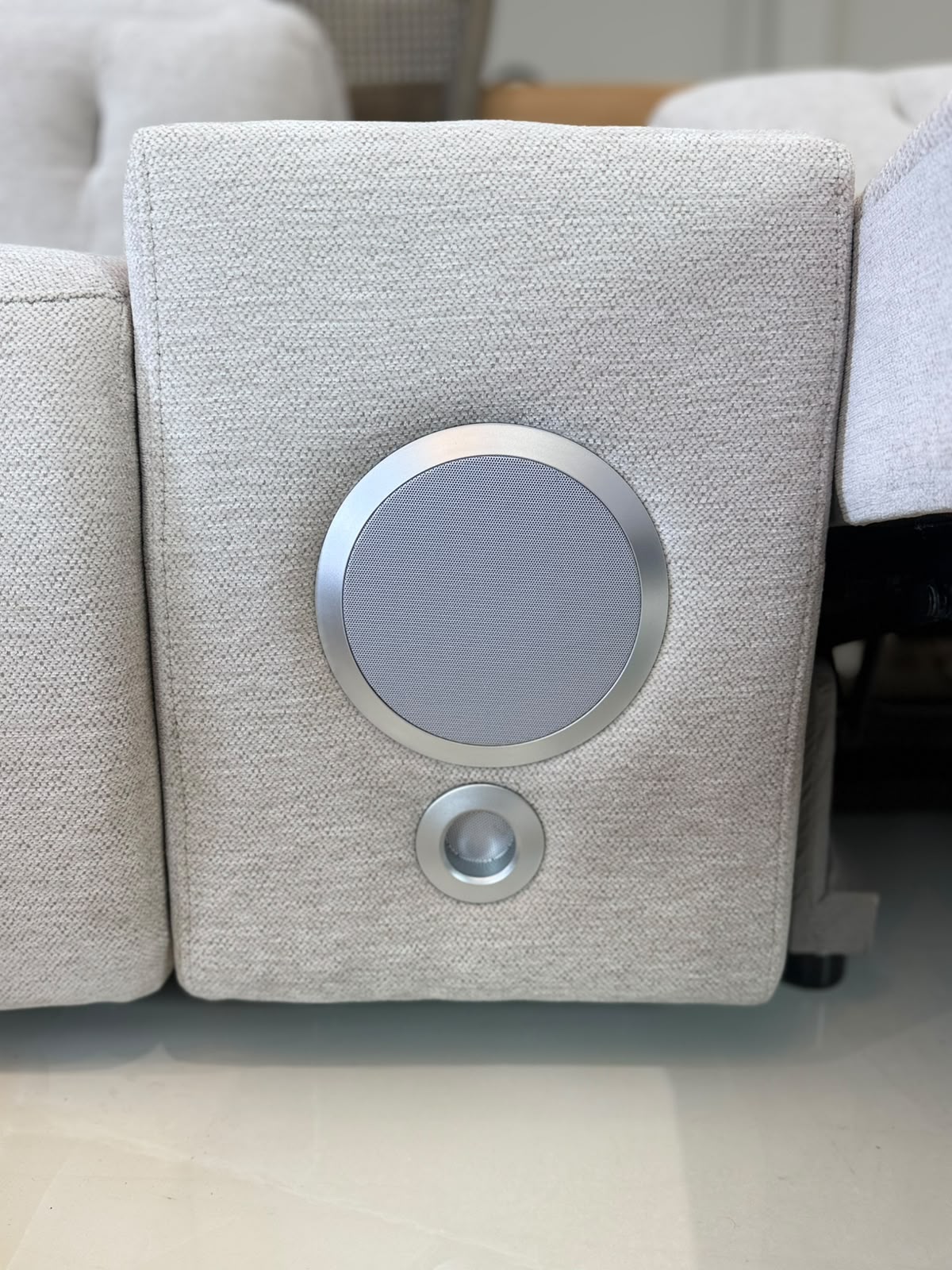 A close-up of a light gray fabric-covered speaker module with a circular silver grille and a smaller circular silver detail below it, mounted on a sofa.