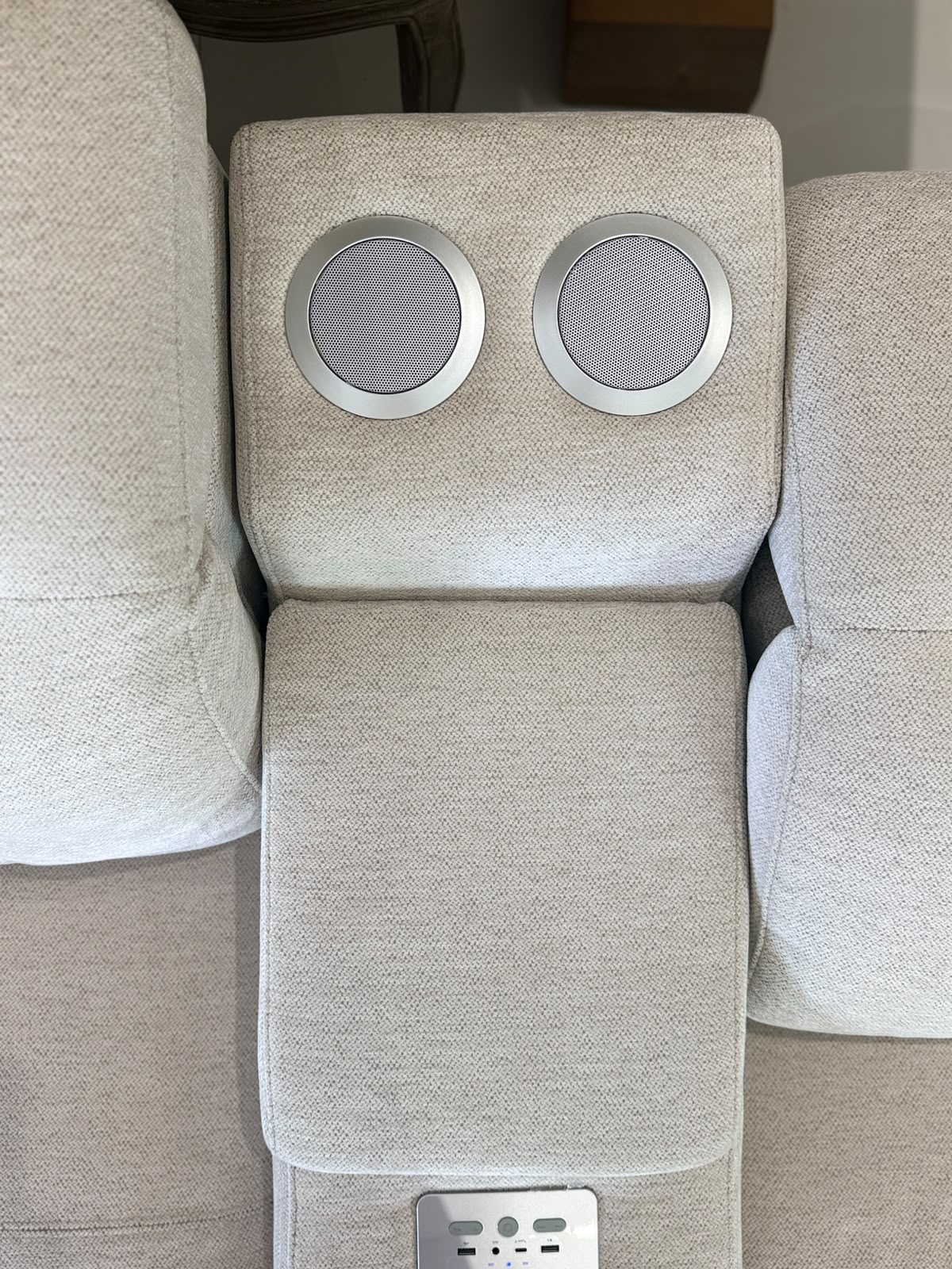 Close-up of a light beige upholstered armrest or center console section with two circular silver speaker grilles on the backrest and a power outlet panel at the bottom.