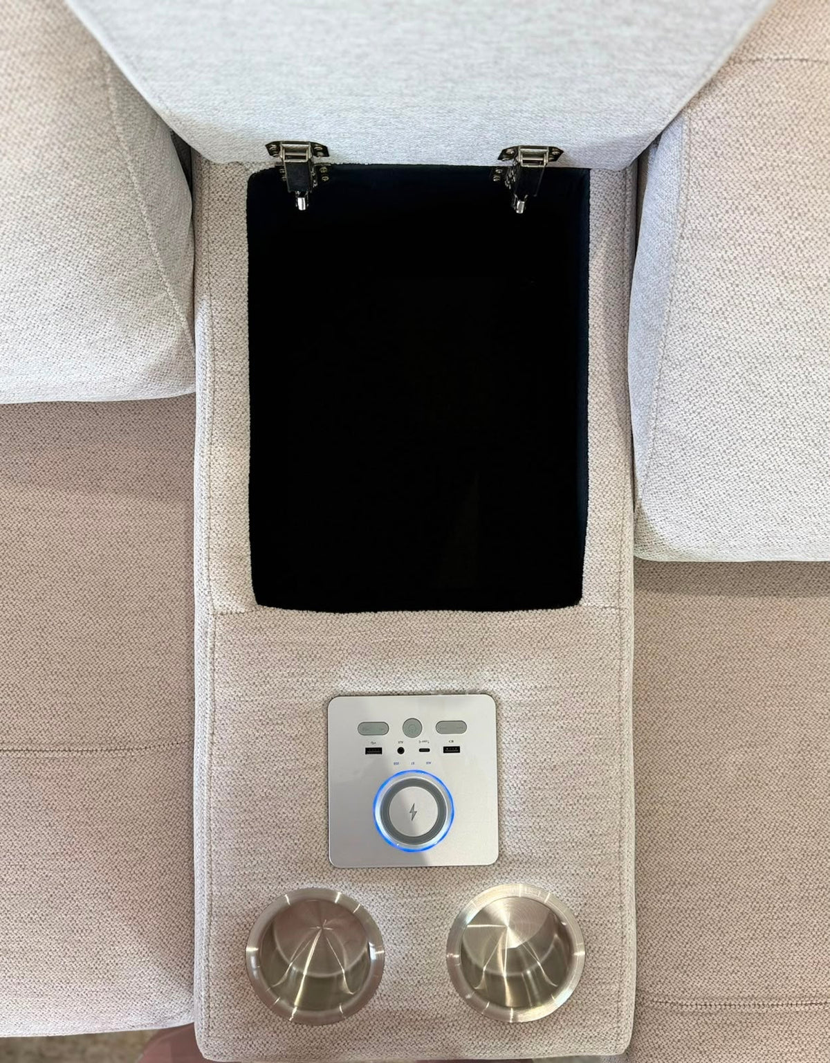 Close-up of a light beige upholstered center console with a black storage pocket, a silver control panel with a blue ring, and two metallic cup holders.
