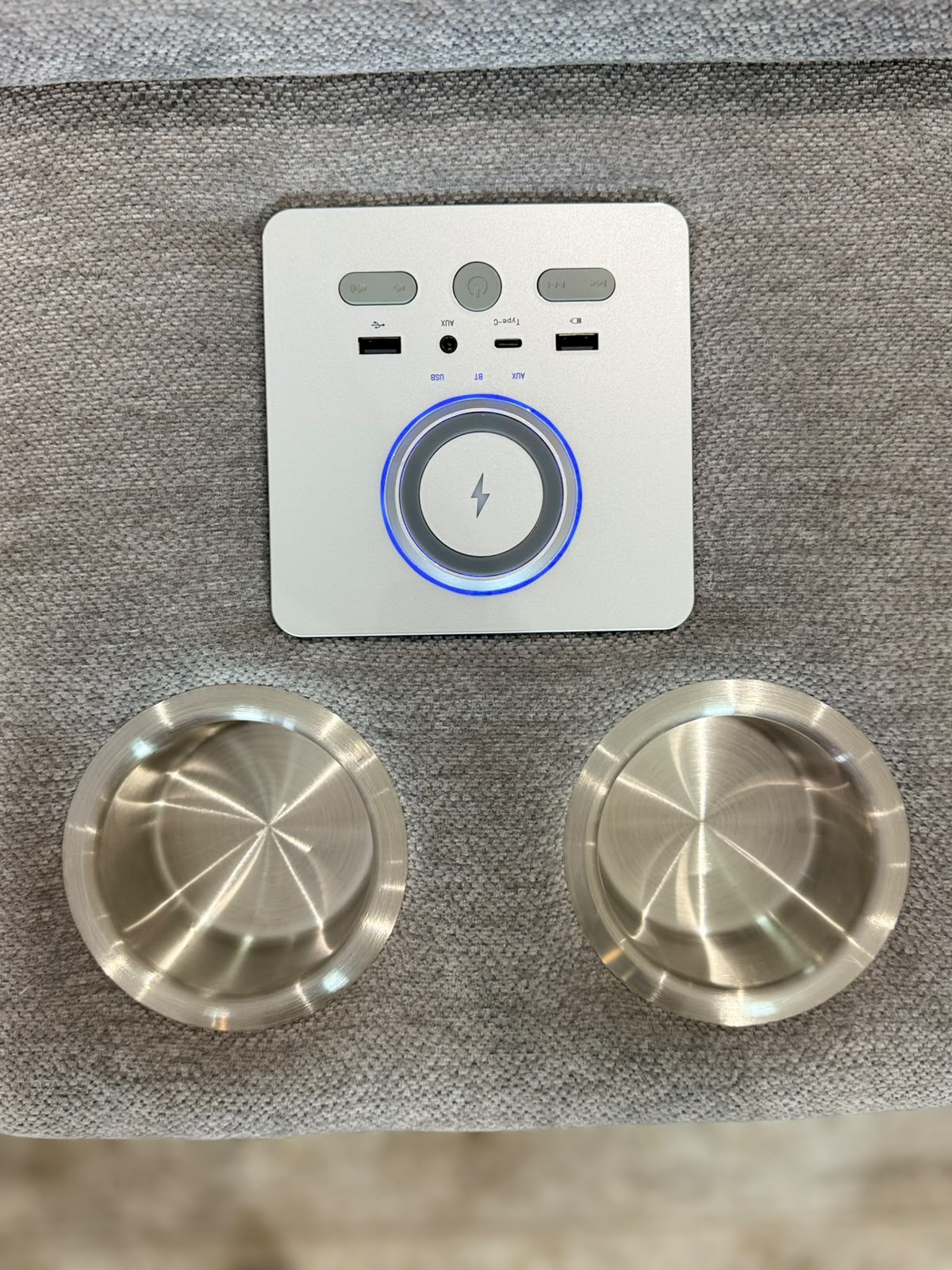 A wireless charging pad set consisting of a white square controller with a blue LED ring and two brushed stainless steel charging cups, displayed on a gray textured fabric surface.