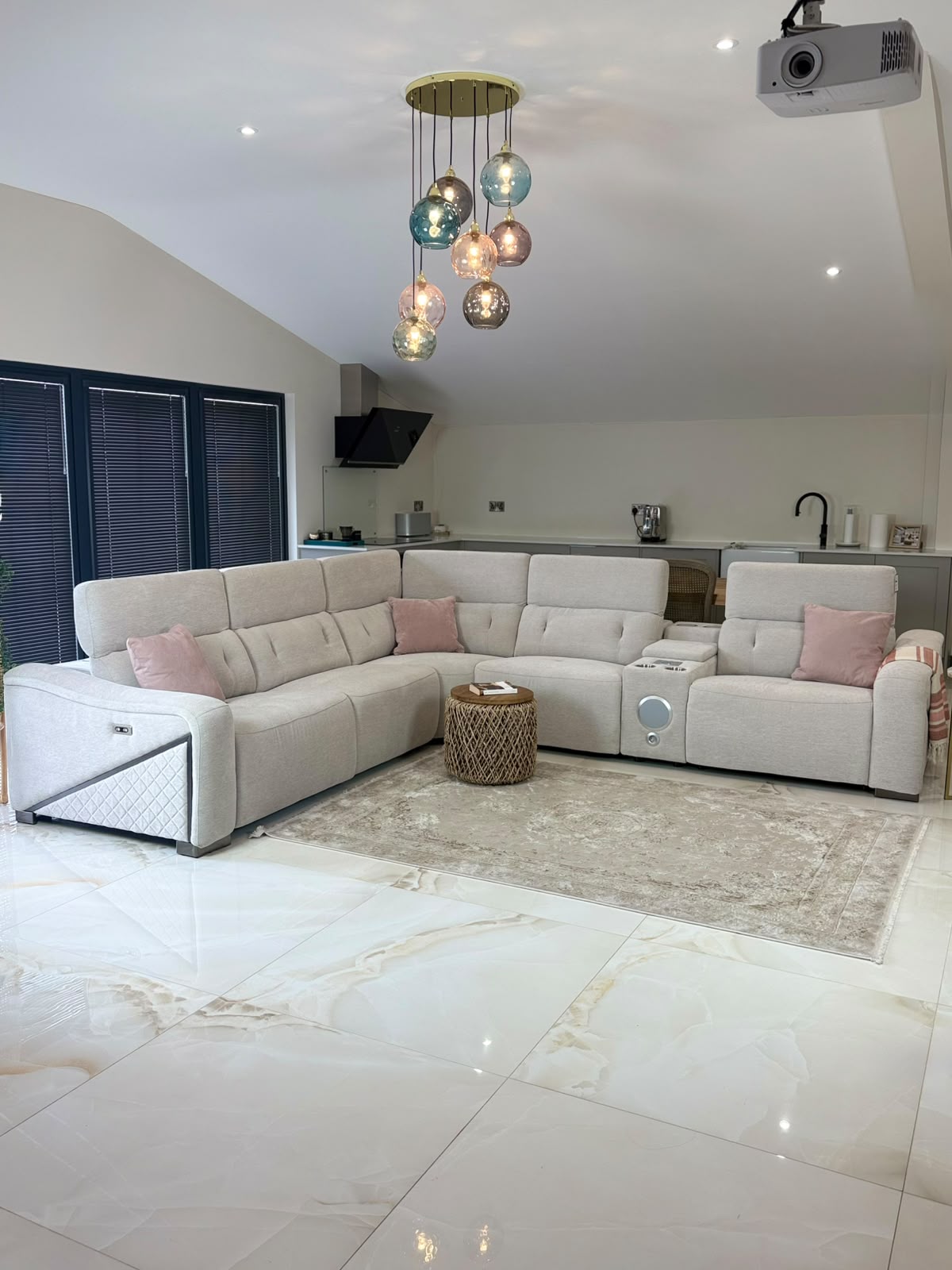 A large cream-colored modular sectional sofa with matching pink accent pillows, set in a modern living room with a patterned rug, a hanging glass chandelier, and a kitchen area in the background.