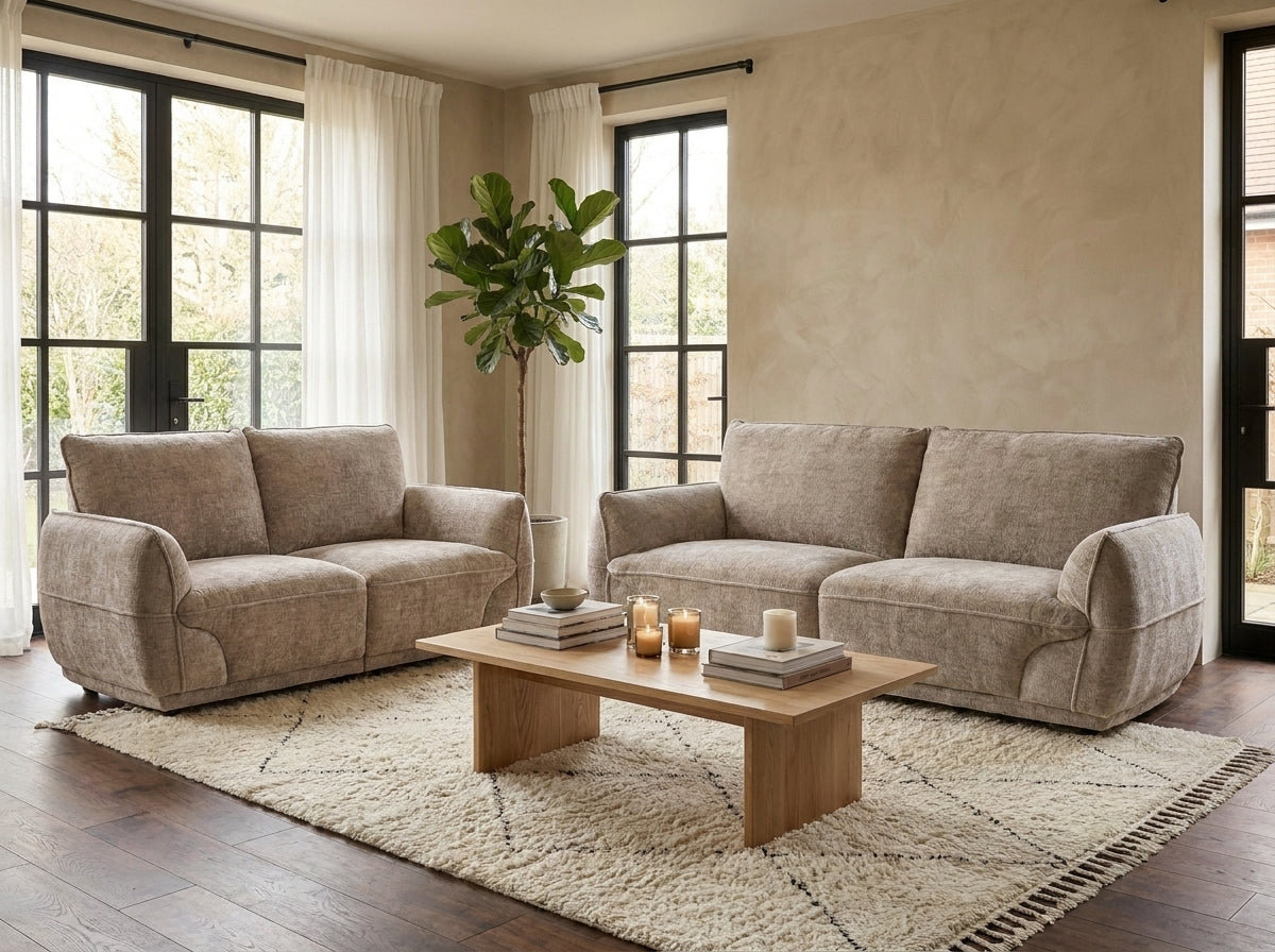 A staged living room featuring a light beige upholstered loveseat and matching sofa arranged around a low wooden coffee table on a large cream rug. The room has warm beige walls, dark wood floors, tall windows with white curtains, and a potted green plant.