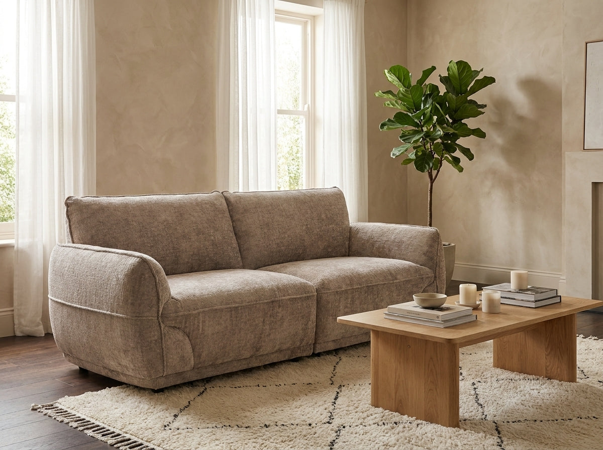 A beige upholstered loveseat sofa in a styled living room setting with a wooden coffee table, textured rug, potted fiddle-leaf fig plant, and warm neutral walls.