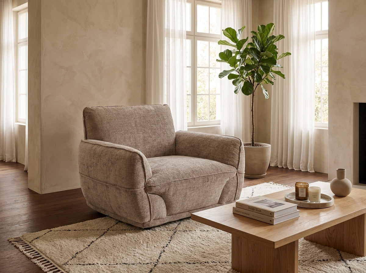 A plush upholstered armchair in a warm taupe-beige fabric, styled in a bright, modern living room with a wooden coffee table, textured rug, and a potted fiddle-leaf fig plant.