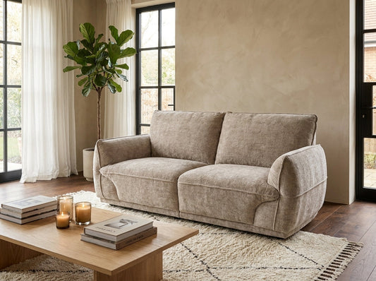 A beige upholstered loveseat sofa in a modern living room setting with a wooden coffee table, textured rug, potted plant, and large windows.