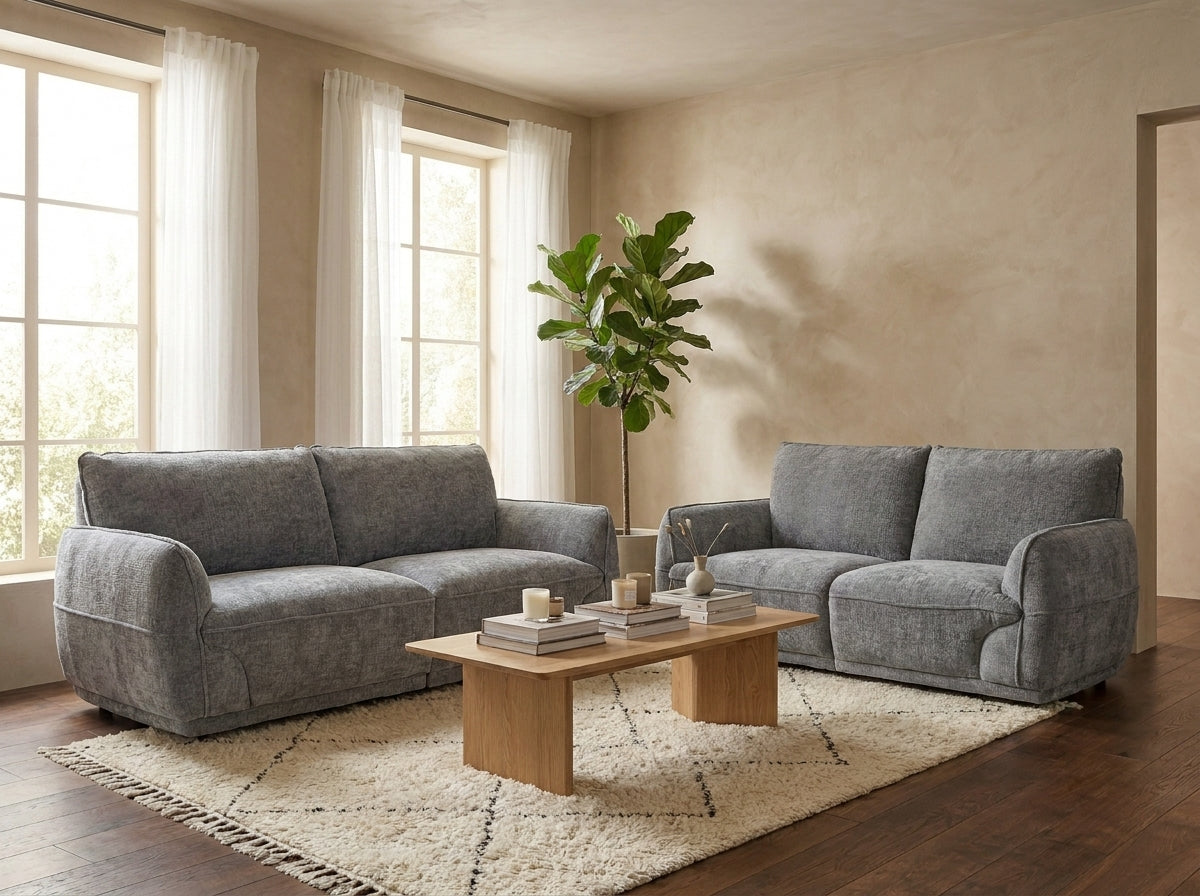 A gray upholstered loveseat and matching armchair arranged in a living room with a wooden coffee table, cream rug, windows with white curtains, and a tall green plant.